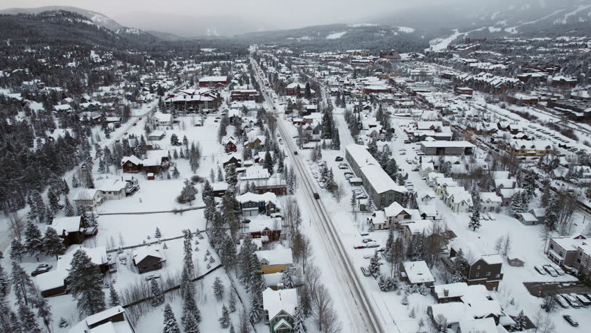 A wide rising aerial of Breckenridge, Colorado, peaceful, beautiful and snow-covered in winter, with cars carrying skis and snowboards into the popular mountain town and world famous resort.