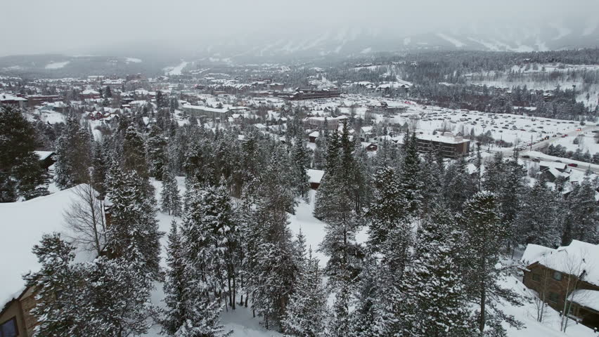 Dynamic aerial flying from the mountains over snow covered fir and evergreen trees revealing Breckenridge, Colorado, a popular mountain town and world famous ski resort for wealthy winter sports.