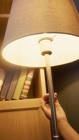 Vertical tilt down shot of bored little Black girl in pyjamas lying in bed under blanket at night in dark room and playing with floor lamp, turning it on and off