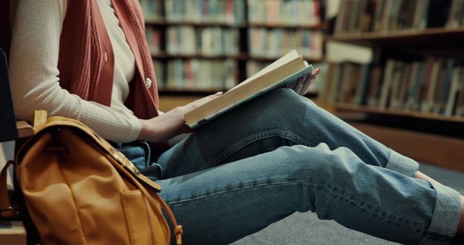 Woman, hands and reading with book in library on floor for education, learning or knowledge. Closeup, female person or student with novel or textbook for story, literature or studying by bookshelf