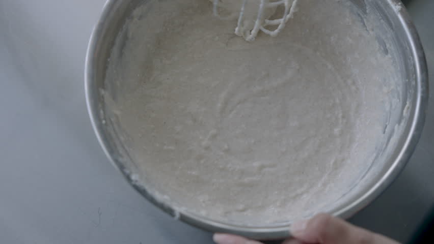 thick bread batter being mixed quickly in metal bowl with balloon whisk while hands hold container during active baking preparation in minimal stainless kitchen setup
