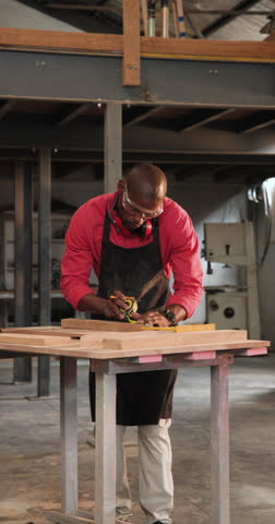 Vertical video: African American mentor measuring board guiding apprentice assembling frame at shop. Craftsmanship, workshop, collaboration, precision, manual, construction, vertical video