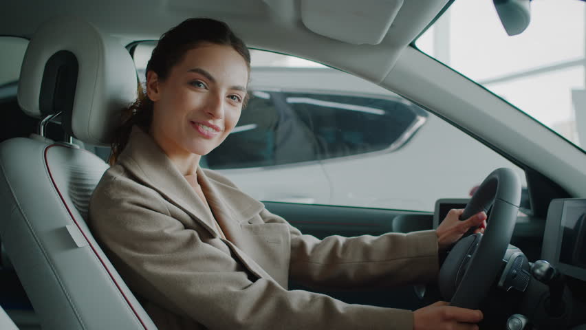 Brunette woman smiles holding steering wheel inside new car in dealership. Confident lady driver satisfied by modern automobile quality in vehicle showroom