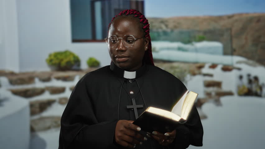 Woman with glasses and red braids wearing clergy attire holds book and points while standing outdoors beside white building with greenery and stone steps nearby indicating conversation.