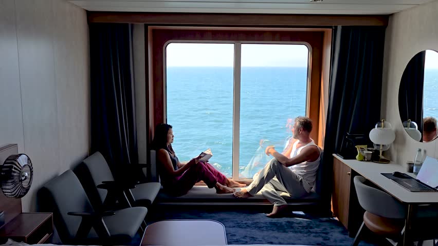 Enjoying a leisurely afternoon beside a large window, a couple relaxes aboard a ship, taking in breathtaking ocean views while reading and unwinding.
