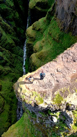 Two adventurers stand on a moss-covered cliff, overlooking a deep ravine with a cascading waterfall. Glymur waterfall canyon in Iceland