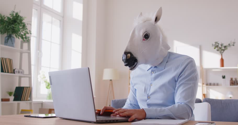 Funny person wearing carnival white horse face mask sitting at desk at home office, waving hand during online video call. Crazy quirky remote worker engaging in virtual communication with colleagues.