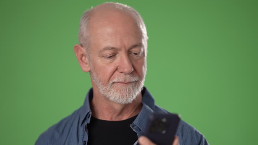 A mature man smiles and waves while using a smartphone, set against a bright green screen. He appears cheerful and connected, enjoying a virtual conversation.