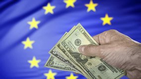 Hand holding some US dollars banknotes in front of a European Union flag, concept of trade war, taxes and tariffs and the 899 Act. - Powered by Shutterstock - Get 15% off with code: PIKWIZARD15