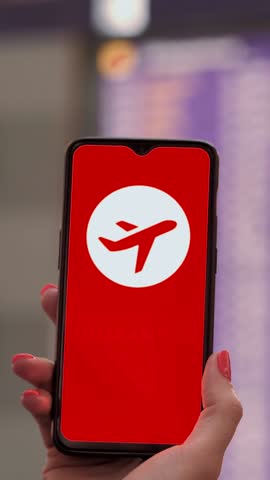 Closeup of hand holding smartphone with airplane icon, quarantine sign, on the screen, in front of flight information board in departures terminal, at the airport.