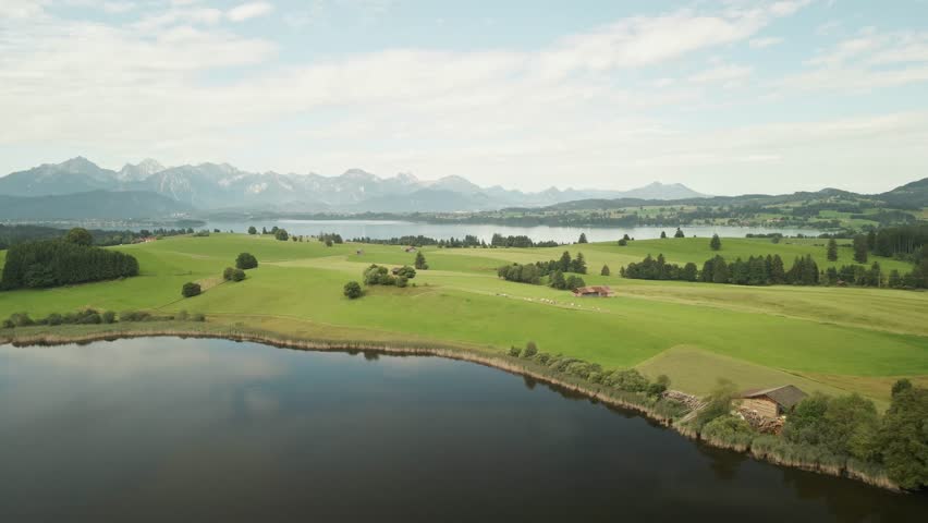 The drone crosses a calm lake in Allgäu, revealing the expansive alpine landscape and lush surroundings.