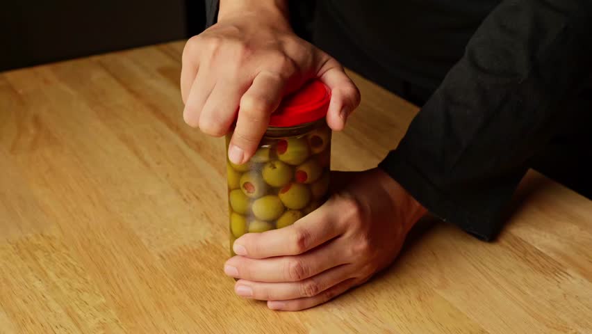 A woman is trying to open a jar of pickle olives with pimento
