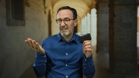 Man speaking in outdoor university corridor holding microphone wearing glasses and blue shirt representing mature hispanic academic public addressing audience enthusiastically. - Powered by Shutterstock - Get 15% off with code: PIKWIZARD15