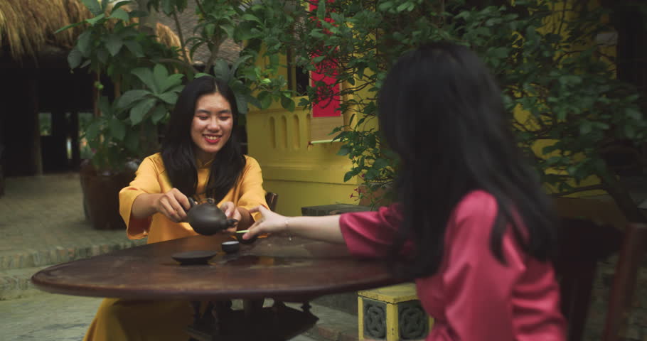 Two Beautiful Asian women chatting while drinking tea. Young tourists in Vietnamese traditional clothes walk through in Vietnam.