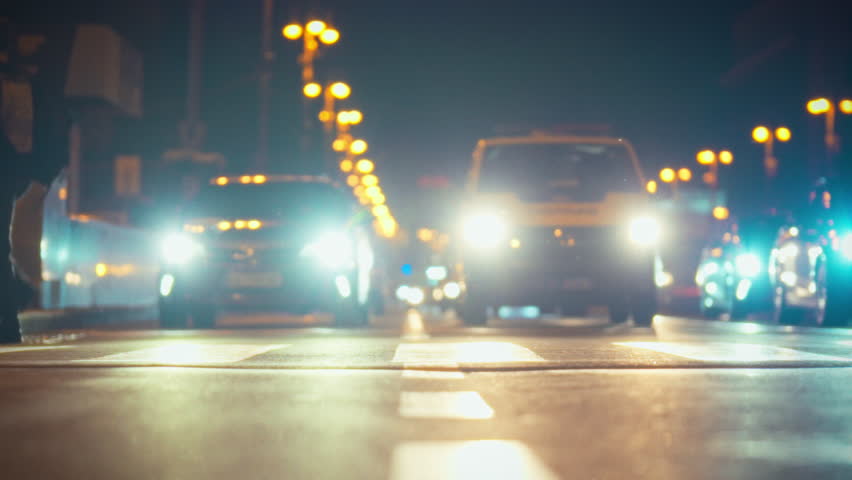 In a vibrant city at night, pedestrians walk across the crosswalk while cars await at a busy intersection. The glow of streetlights and headlights creates a lively atmosphere