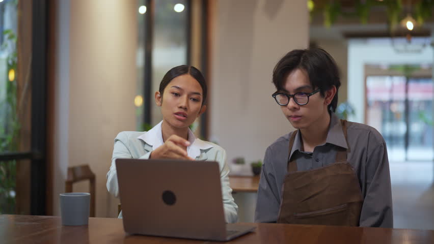 Young professional woman mentoring a barista in front of a laptop at a coffee shop. Female business coach or consultant teaching small business operations, technology usage.