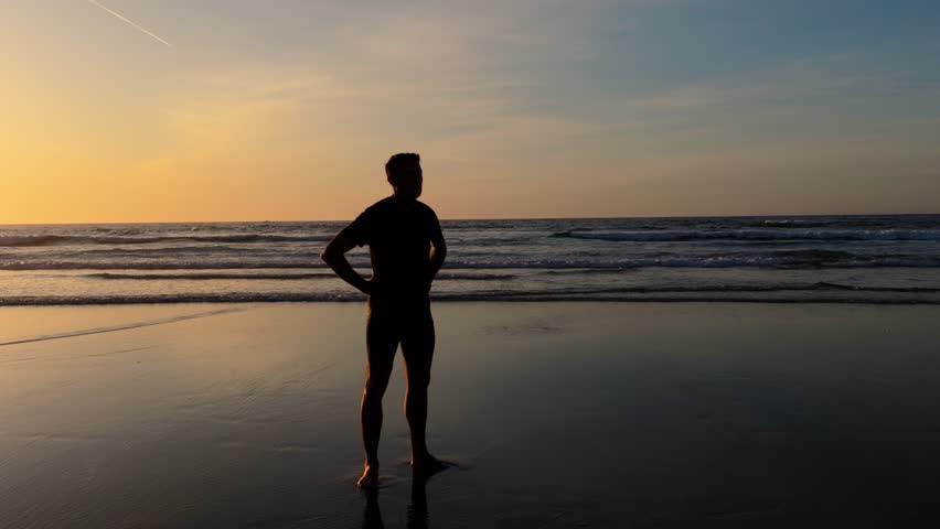 Silhouette Of A Man Standing On Beach At Sunset, With Golden Sun And Waves In Background. orbiting shot