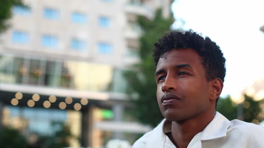 Young man standing in urban city center, embodying hope and aspirational dreams while gazing across blurred architectural landscape during evening light
