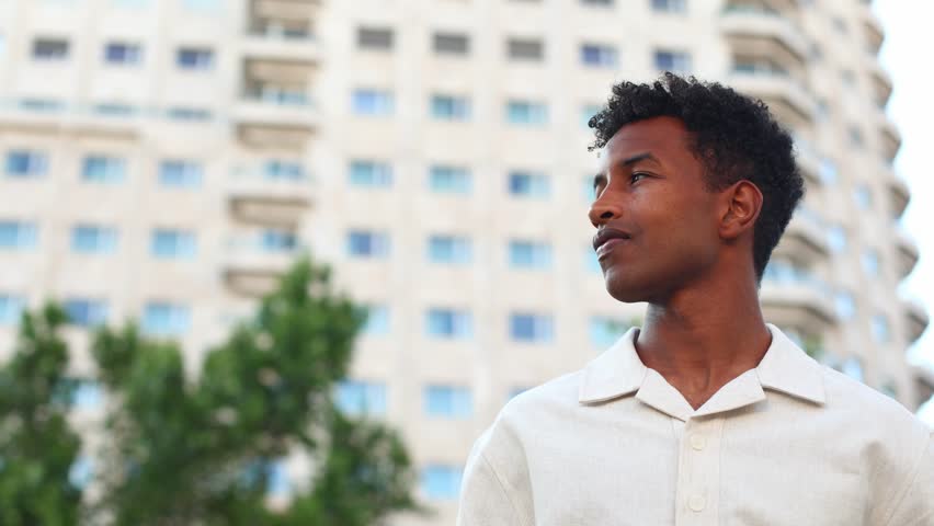 Pensive young professional standing amid urban landscape, gazing upward with contemplative expression while surrounded by architectural structures and city environment