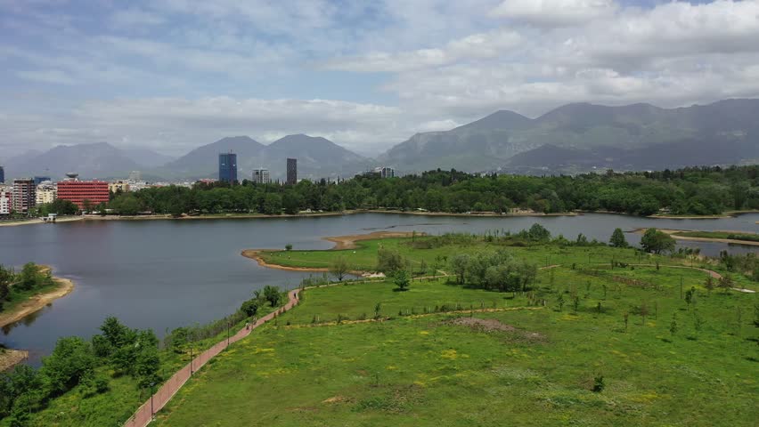 Forward drone shot over Tirana’s Artificial Lake in Albania’s National Park, revealing lush greenery, calm waters, city skyline, and mountains in the background under a moody, cloudy sky.