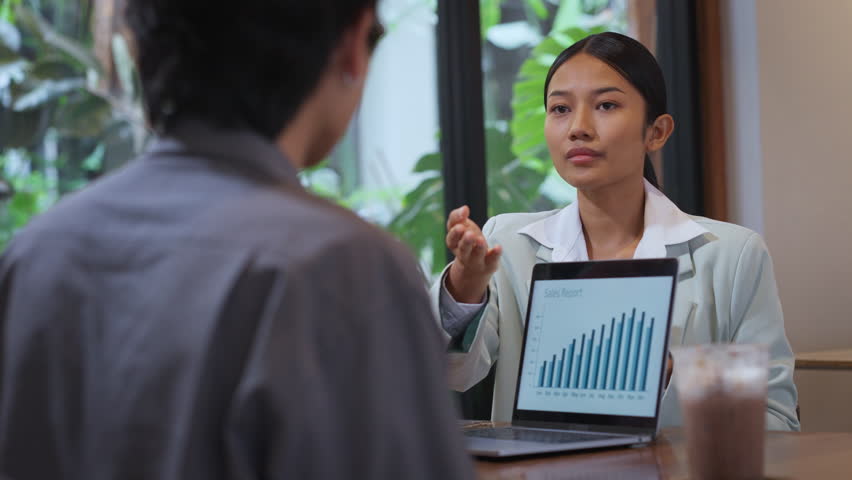 Confident Asian businesswoman presenting a marketing plan using a laptop during a business meeting in a casual café setting. Team collaboration, project strategy, and modern workplace concept.