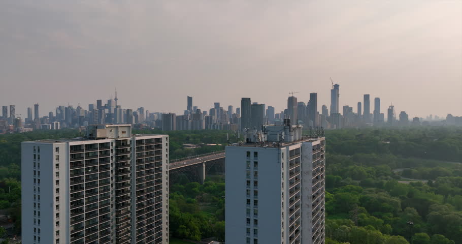 Aerial tracking shot with apartment buildings in the foreground reveals the downtown Toronto skyline, Iconic Tower, and Bloor Viaduct over the lush Don Valley Parkway during a hazy day.
