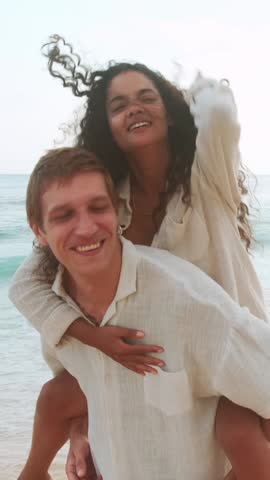 A diverse couple shares a joyful moment at the beach, with one partner giving the other a piggyback ride.