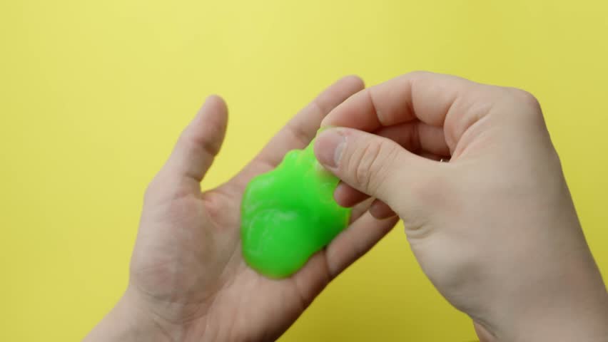 Hands stretching and pressing a wavy green slime toy for ASMR and stress relief against a yellow background, highlighting texture and movement, static camera, real time, close-up view