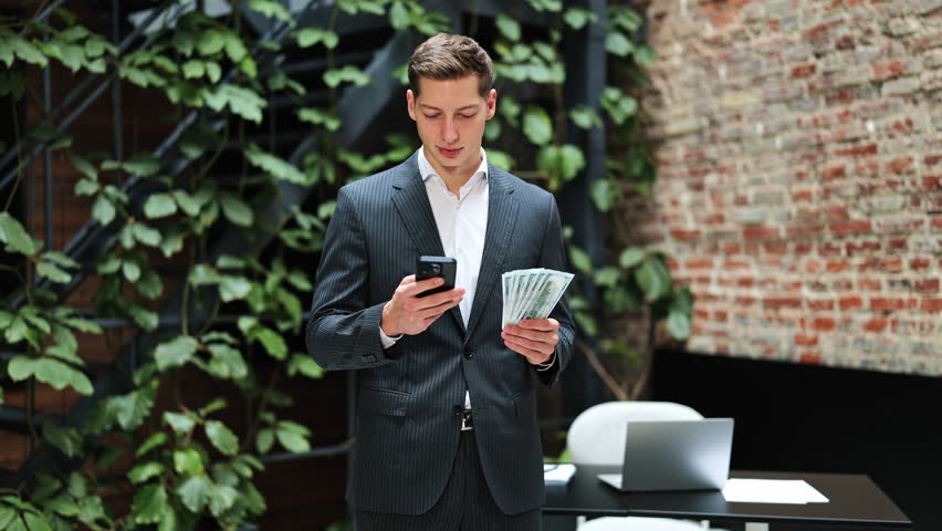 Cheerful businessman in suit holds cash and uses phone in modern office. He feels excited about his income.