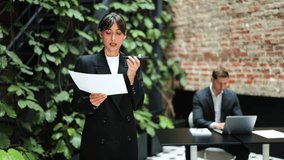 Confident businesswoman in a black suit records a voice message in a modern office. Businessman works on laptop on background. - Powered by Shutterstock - Get 15% off with code: PIKWIZARD15