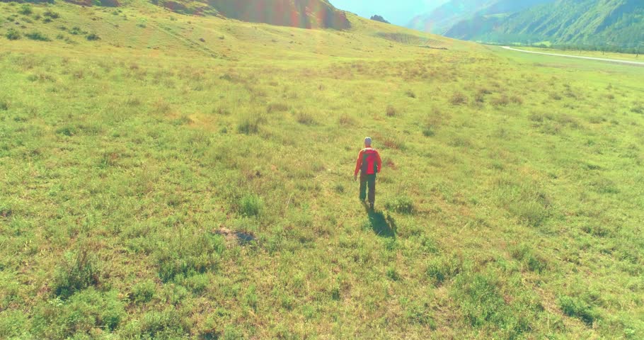 Aerial 4K UHD view. Low altitude flight over young man tourist walking across green mountain field. Huge rural valley and sunny meadow at summer day. Backpack hiking guy.