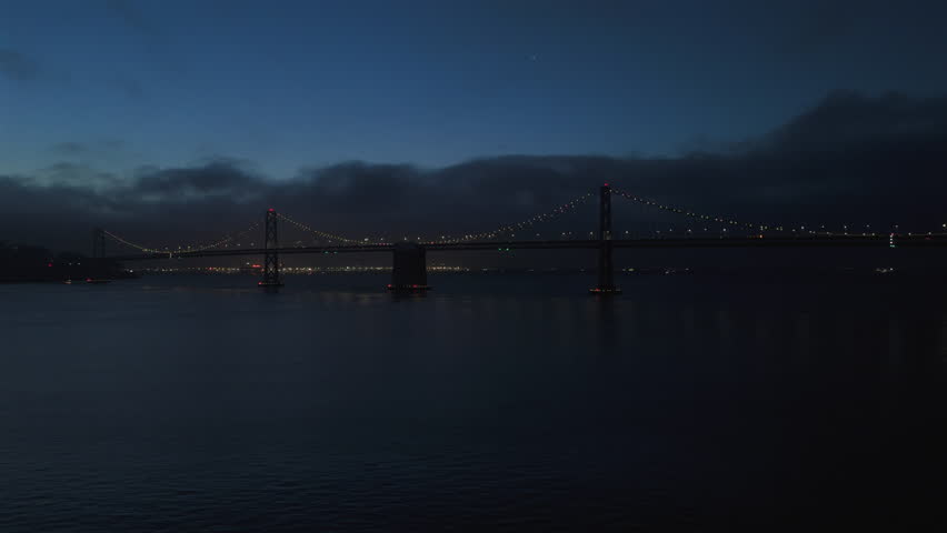 Breathtaking aerial shot of illuminated Bay bridge in San Francisco spanning over calm, reflective waters at twilight, beautifully showcasing serene cityscape and car traffic on the bridge 