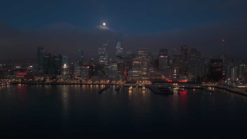 Stunning aerial shot of San Francisco skyline captured beautifully at night with full moon shining, vibrant city lights illuminating the skyline, while iconic fog gracefully rolls in over the bay area