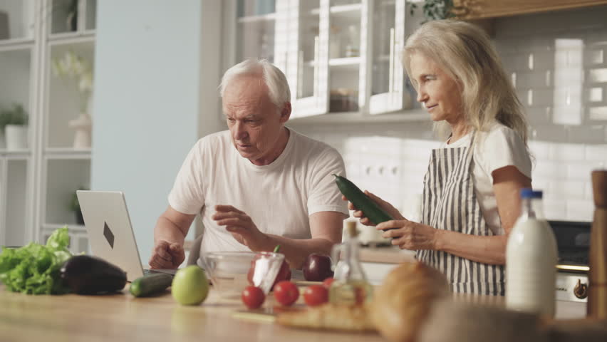 Elderly people cooking at home, old man and woman looking for video recipe. Elderly man surfing internet with cook web-sites and mature wife cooking vegetable salad, healthy nutrition and organic food - Powered by Shutterstock - Get 15% off with code: PIKWIZARD15