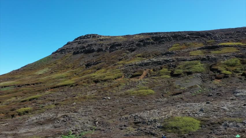 Drone panning shot of Icelandic fjord with snow-capped mountains, blue water, and colorful coastal terrain. Bright sky and wide open space highlight beauty of natural untouched landscape.