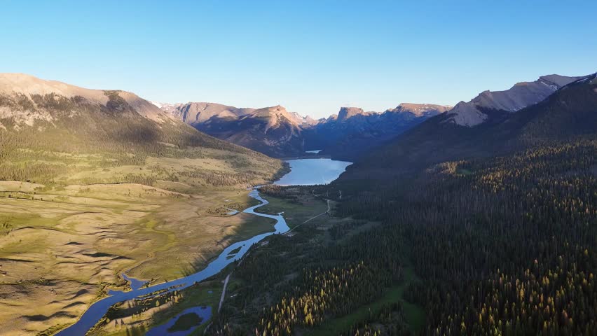 Aerial drone shows winding river flowing through green valley into large lake between tall forest and rocky mountain in scenic peaceful West Virginia landscape under clear blue sky in daylight