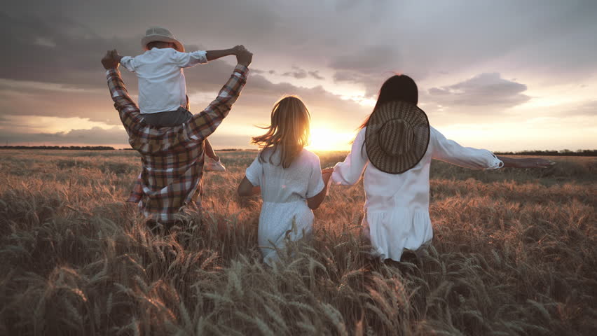 Happy family running to sunrise over golden rye field, holding hands, rejoicing. Back view of parents and children admiring fascinating sunset above agricultural field, childhood and parenthood moment