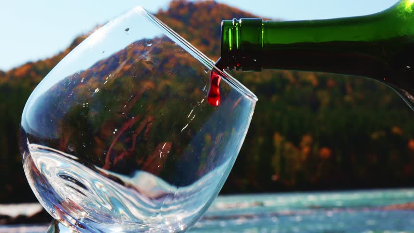 Pouring rich red wine into a glass with a picturesque river and vibrant autumn trees in the background during a sunny day. Experience the tranquility of nature.