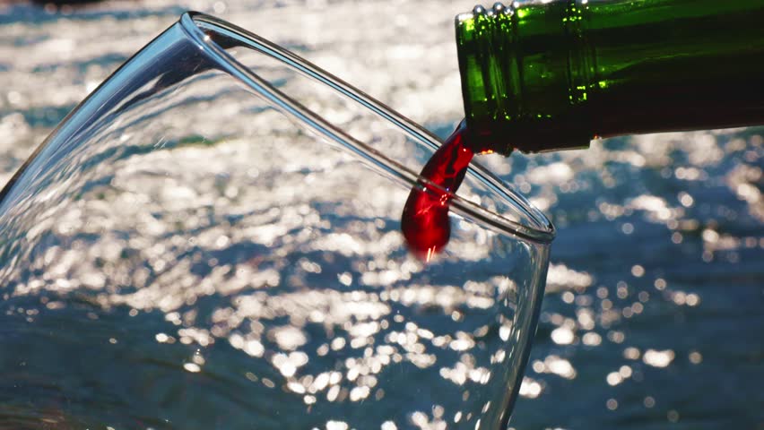 A bottle of red wine is being poured into a transparent glass, catching the light from the sun. The background features shimmering water, creating a refreshing atmosphere.