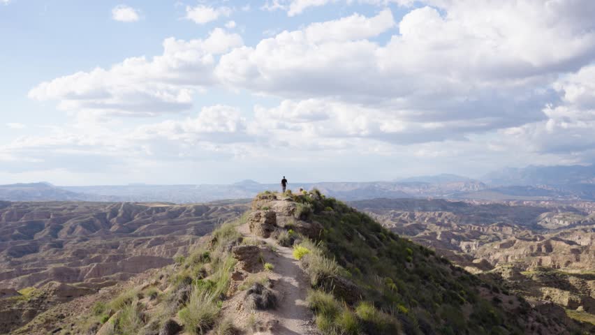 Distant shot of man walking along narrow ridge trail surrounded by vast rugged hills under cloudy blue sky capturing remote scenic view in Gorafe desert, Granada, Spain