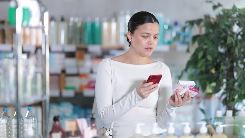 Interested young Asian female customer scanning different paracetamol packages with phone to compare prices, ingredients, and reviews online, looking for painkiller or fever reducer in pharmacy 