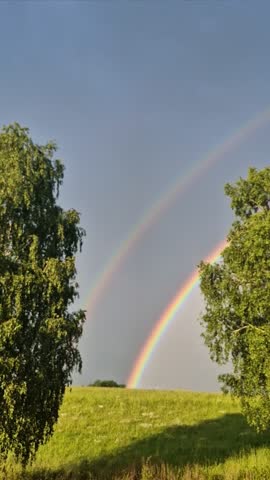 A double rainbow playing with all colors, arching over the field and deciduous trees in the bright light of the setting sun, creating the impression of a peaceful and magical atmosphere over the Czech