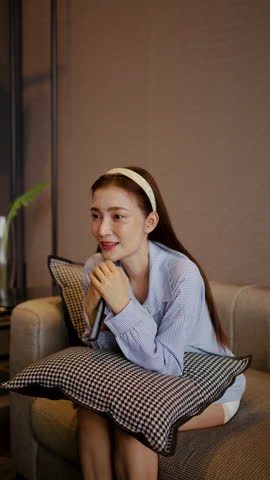 Relaxed adult asian woman sits comfortably on her living room sofa, engrossed in her favorite TV show or movie with remote in hand, as the evening unfolds peacefully.