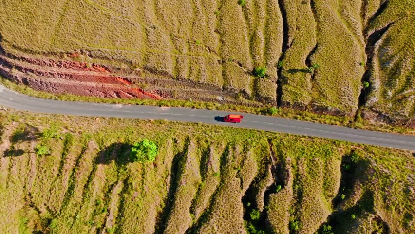Aerial view of a red Taxi Brousse navigating a winding mountain road through the dramatic and colorful terrain of Madagascar. Perfect for transport, travel, and landscape themes.