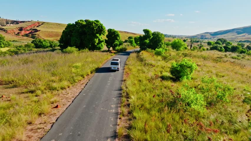Aerial view of a white bus driving along a remote road through grassy highlands and green trees in Madagascar. Peaceful rural transportation scene under a clear sky.