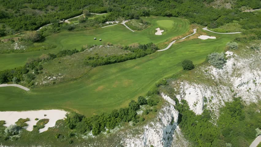 Scenic aerial view of a coastal golf course with green fairways, sand bunkers, golf carts, and players, surrounded by lush vegetation and turquoise sea