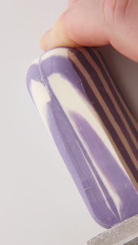 Purple and white striped soap being cut with metal knife creating uniform slices as hand guides cutting motion producing calming ASMR sounds and satisfying visual patterns on surface