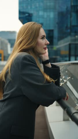 Profile of thoughtful and confident businesswoman in a dark suit stands on a modern office terrace, leaning on a railing - dolly in, slow motion