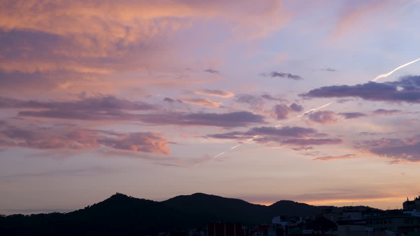 A horizontal panning shot of a vibrant sunset over the city of Mataró, Spain. Colorful clouds, mountain silhouettes, and urban rooftops create a peaceful scenic view.