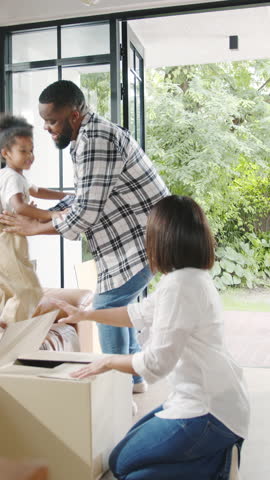 Vertical Screen : Happy African American young family bought new house. Mom, Dad, and child happy hold cardboard boxes for move object and having fun laugh into modern home. New real estate dwelling.